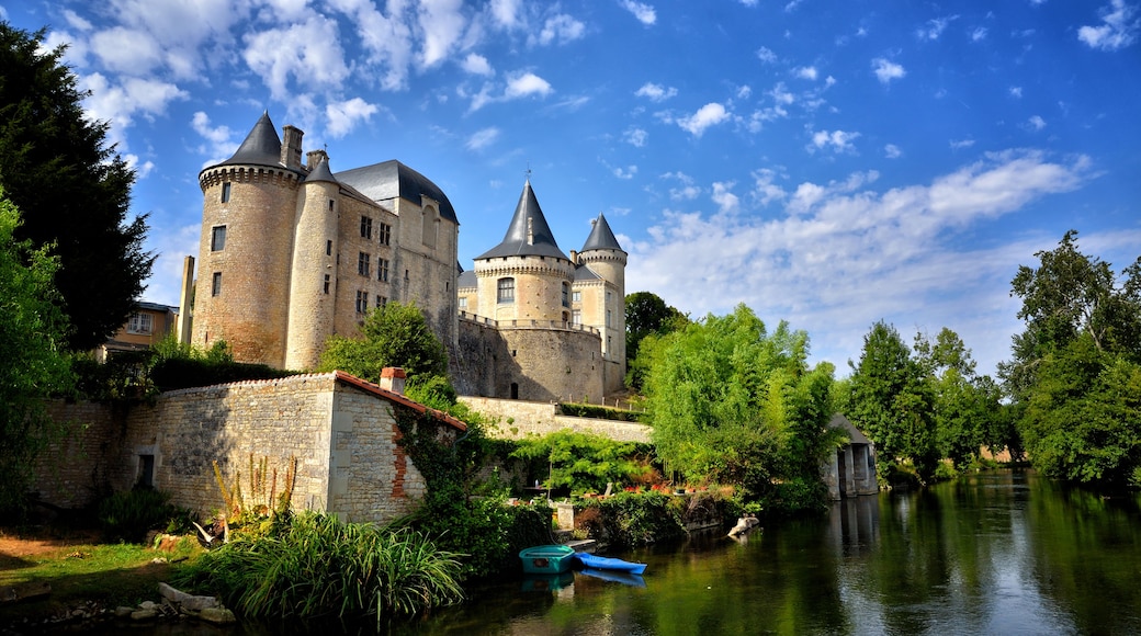 Un château historique doté de nombreuses tours, une rivière au premier plan et une végétation luxuriante qui entoure l’édifice.