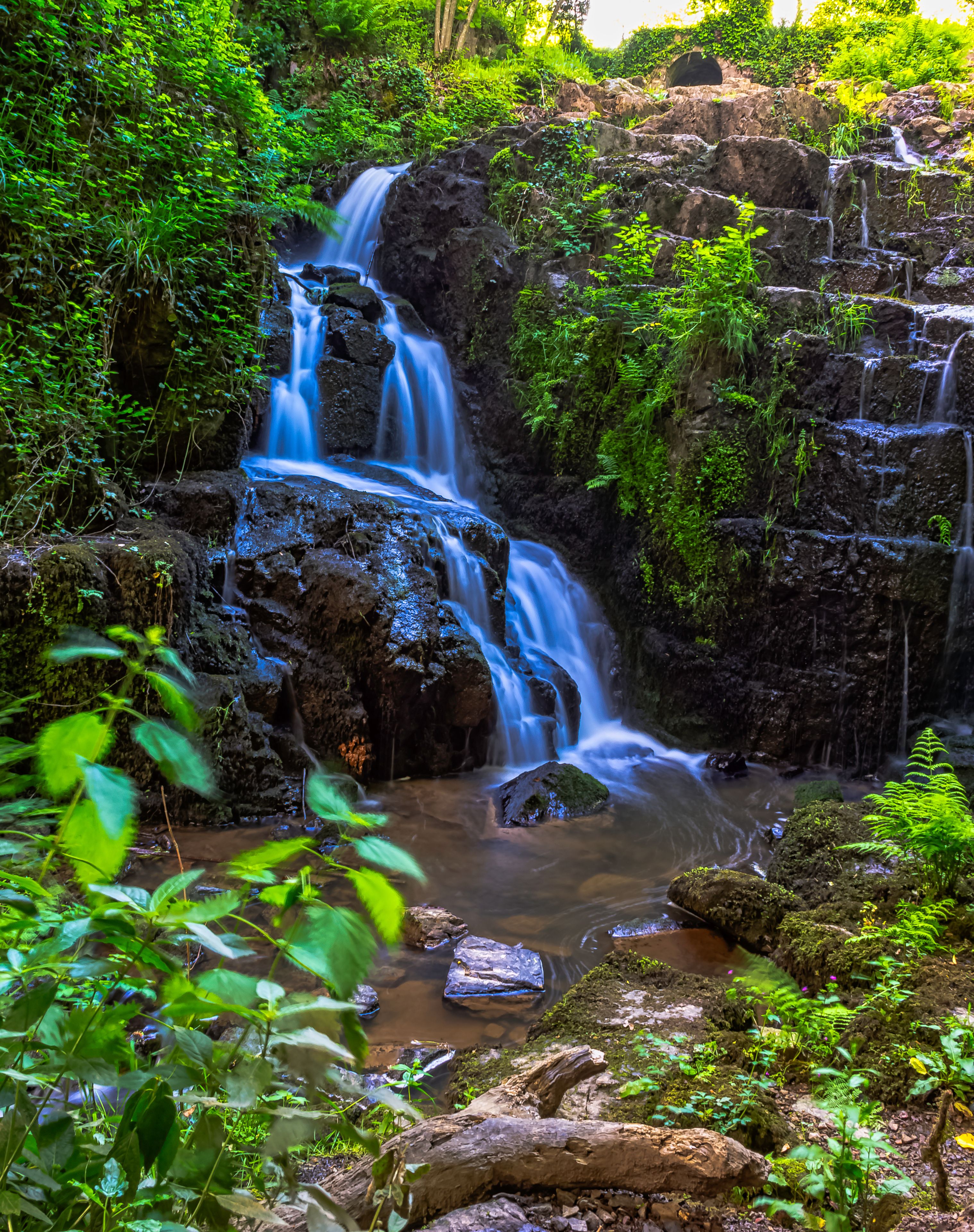 A multi-tiered waterfall surrounded by lush greenery and moss-covered rocks.