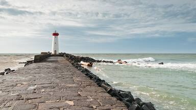 A lighthouse on a stone pier extending into the sea, with waves crashing against the rocks.