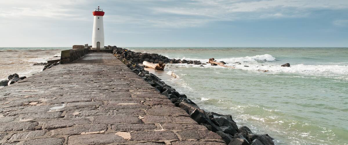 A lighthouse on a stone pier extending into the sea, with waves crashing against the rocks.