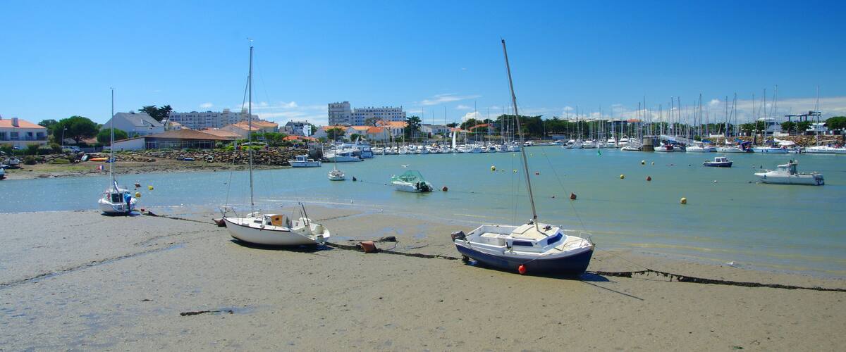 A beach with several boats anchored near the shore, a clear blue sky, and buildings in the background.