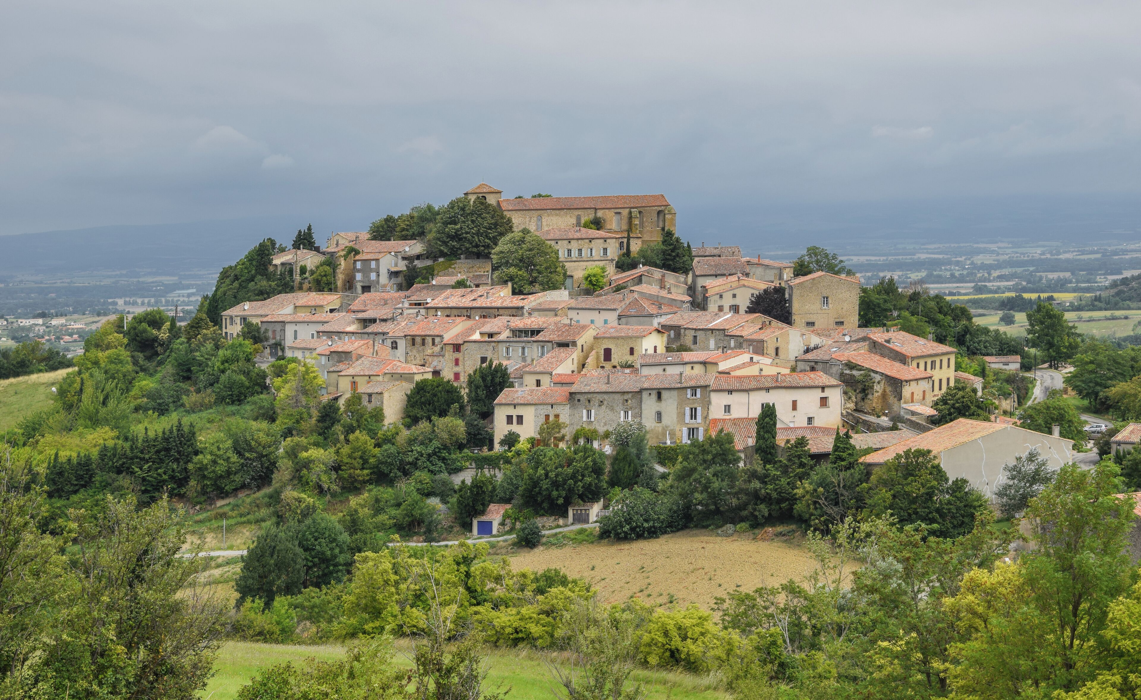 A hillside village with terracotta-roofed buildings, surrounded by greenery and a distant view of the countryside.