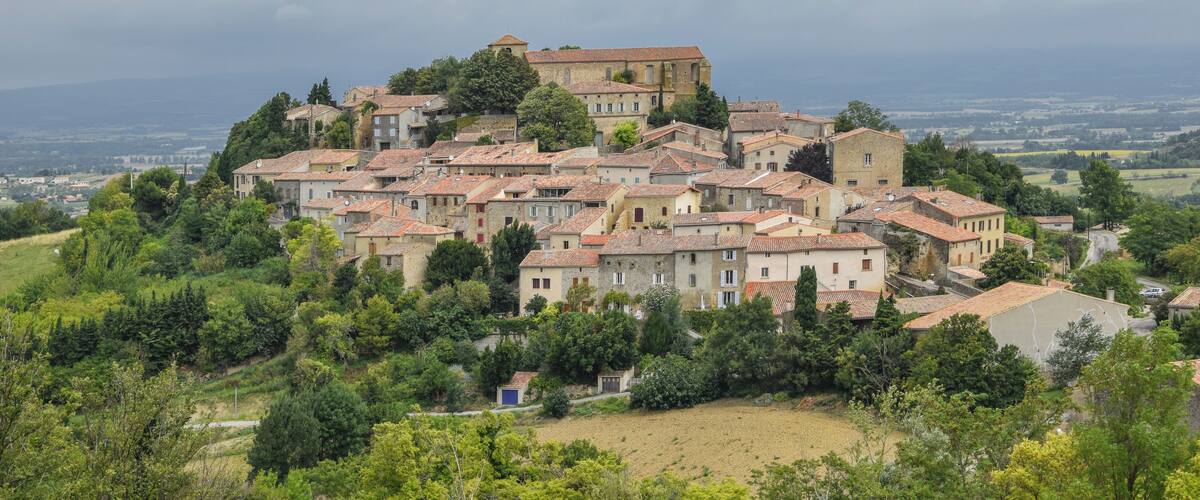 A hillside village with terracotta-roofed buildings, surrounded by greenery and a distant view of the countryside.