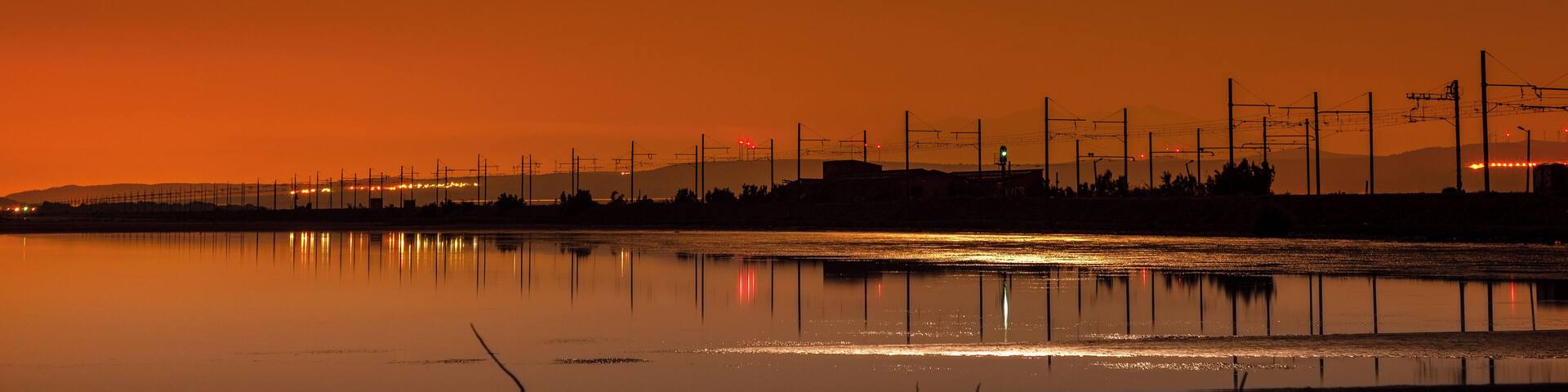 A sunset over a calm water body with a silhouette of a distant city skyline and power lines.