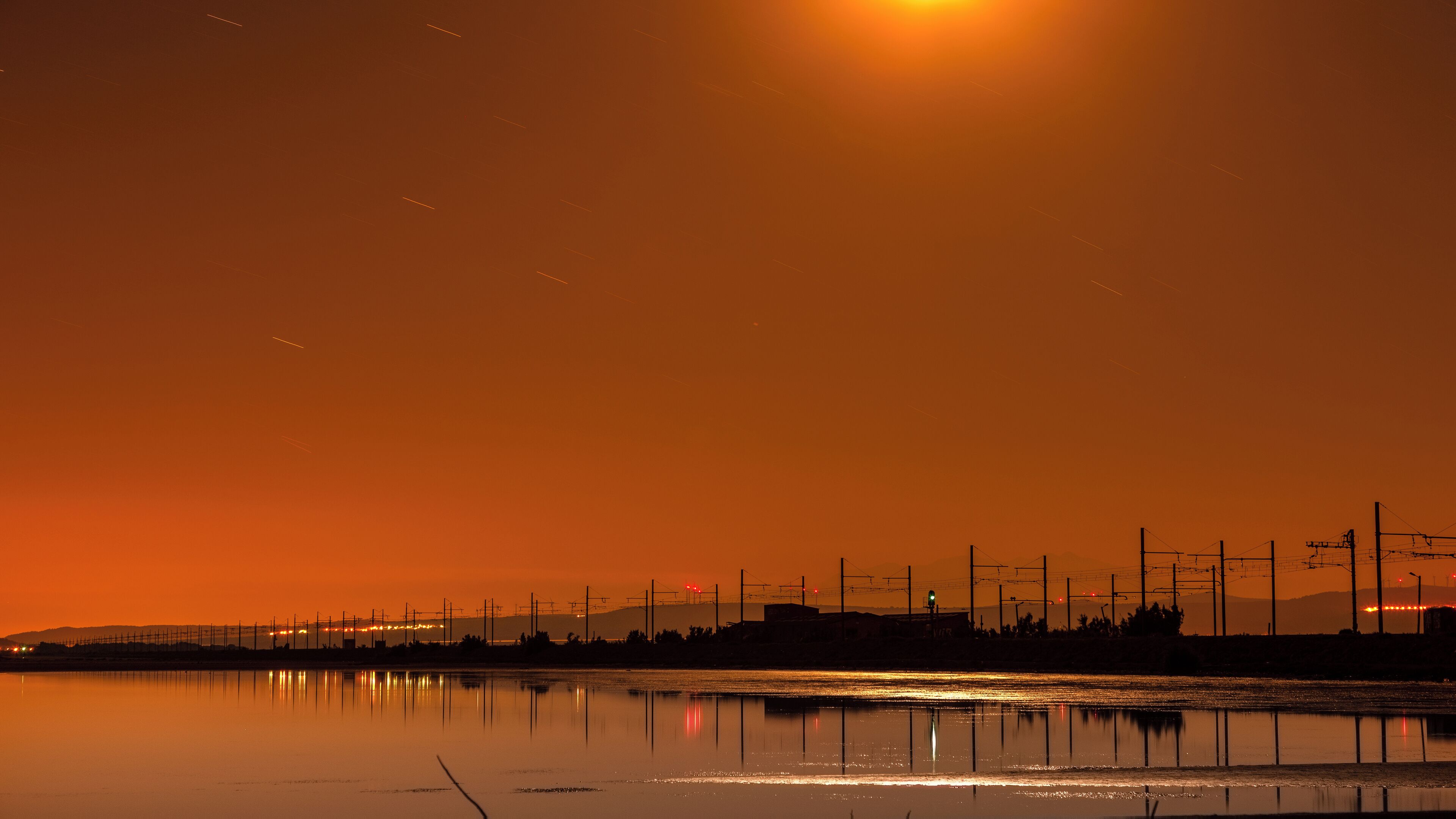 A sunset over a calm water body with a silhouette of a distant city skyline and power lines.
