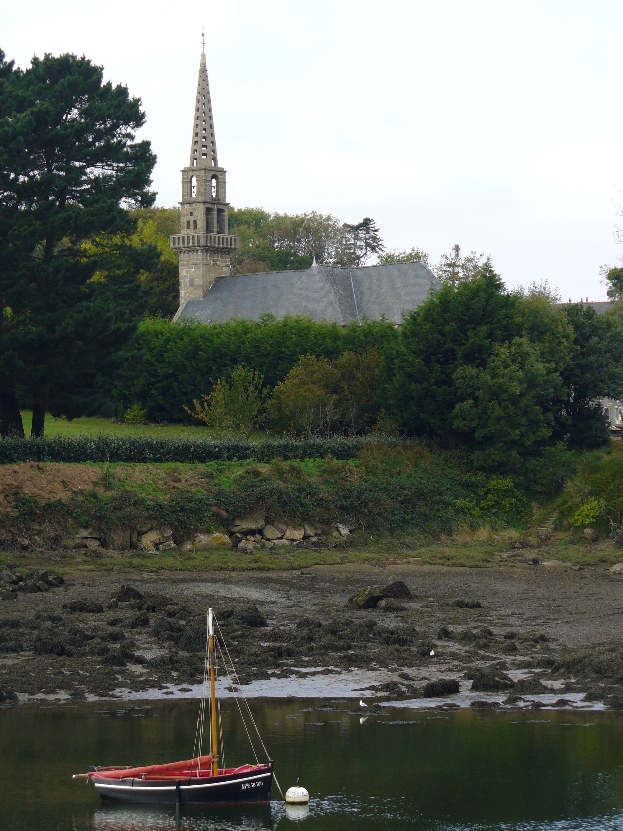 A boat on a calm body of water, a church with a tall spire, and dense greenery in the background.