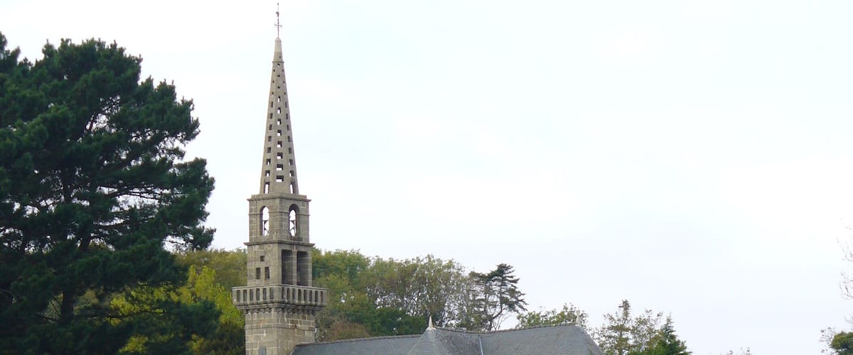 A boat on a calm body of water, a church with a tall spire, and dense greenery in the background.