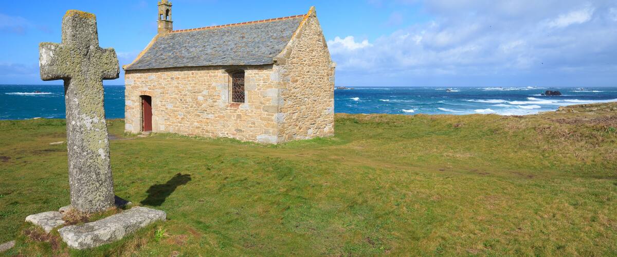 A stone church with a cross in front, a grassy field, and the ocean in the background.