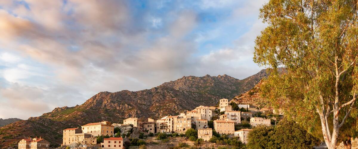 A coastal village with stone buildings, a winding road, and mountains in the background.