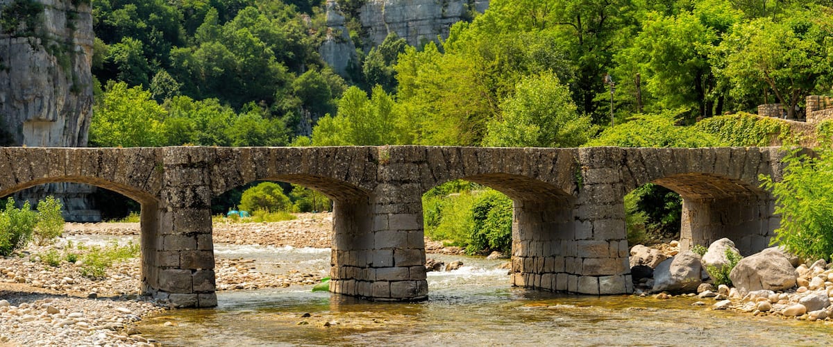 A stone bridge over a river with rocky banks and greenery in the background.