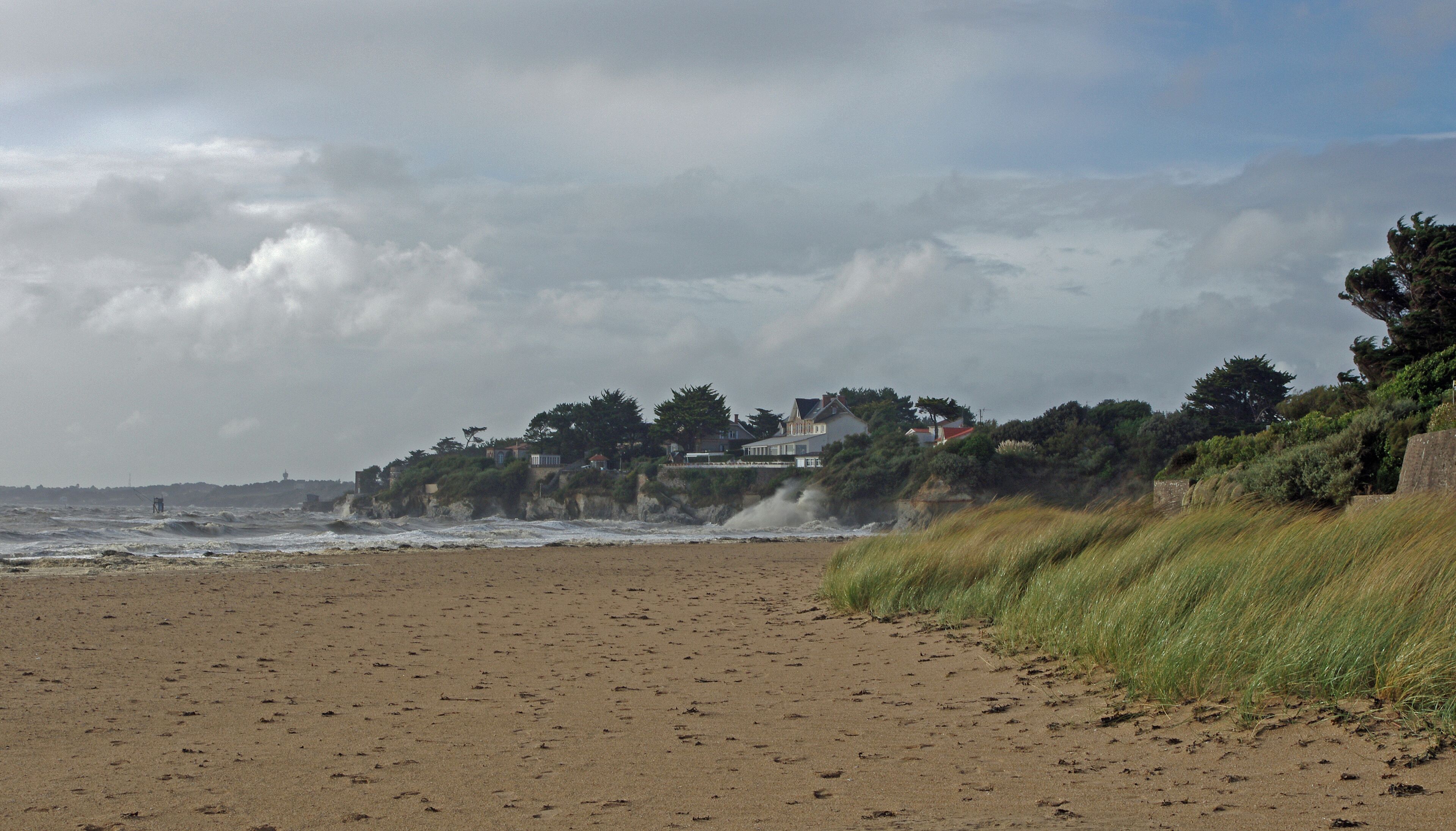 A sandy beach with waves crashing onto the shore, a grassy cliff, and houses on a hillside.