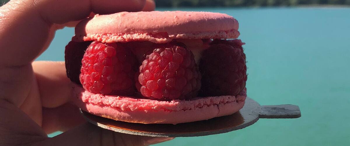 A hand holding a raspberry macaron with raspberries on a spoon over a turquoise lake with mountains in the background.