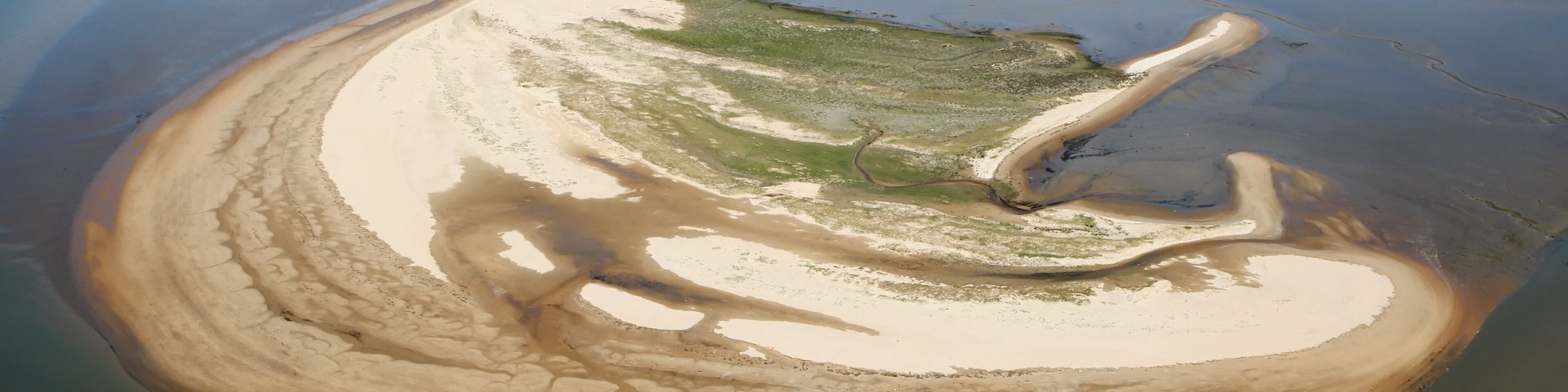 Vista aérea de un paisaje costero con una playa arenosa, una bahía curva y un pequeño asentamiento a lo lejos.