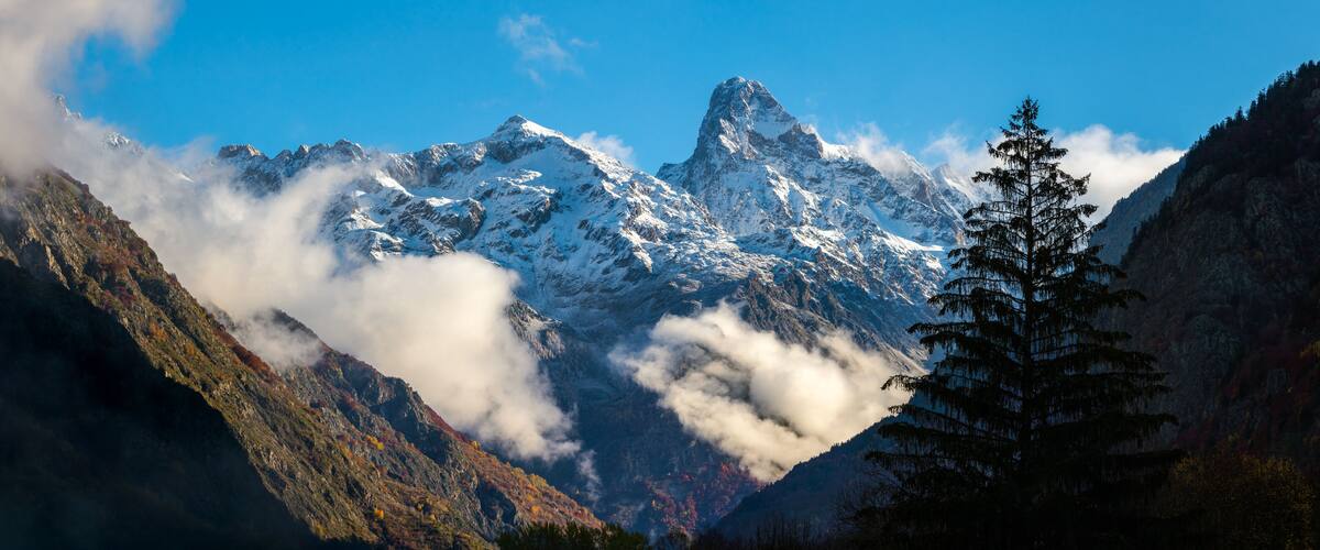 A snow-capped mountain range with a prominent peak, surrounded by clouds and trees.