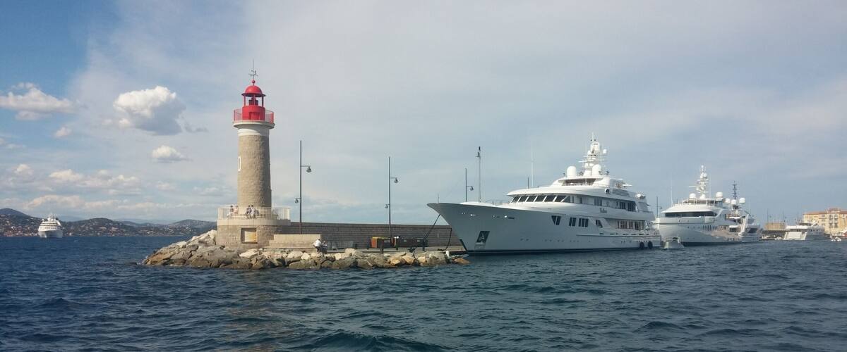 A lighthouse, a large yacht, and a smaller boat docked near a rocky shore.