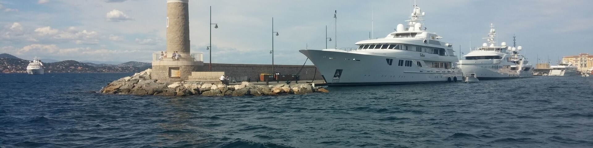 A lighthouse, a large yacht, and a smaller boat docked near a rocky shore.