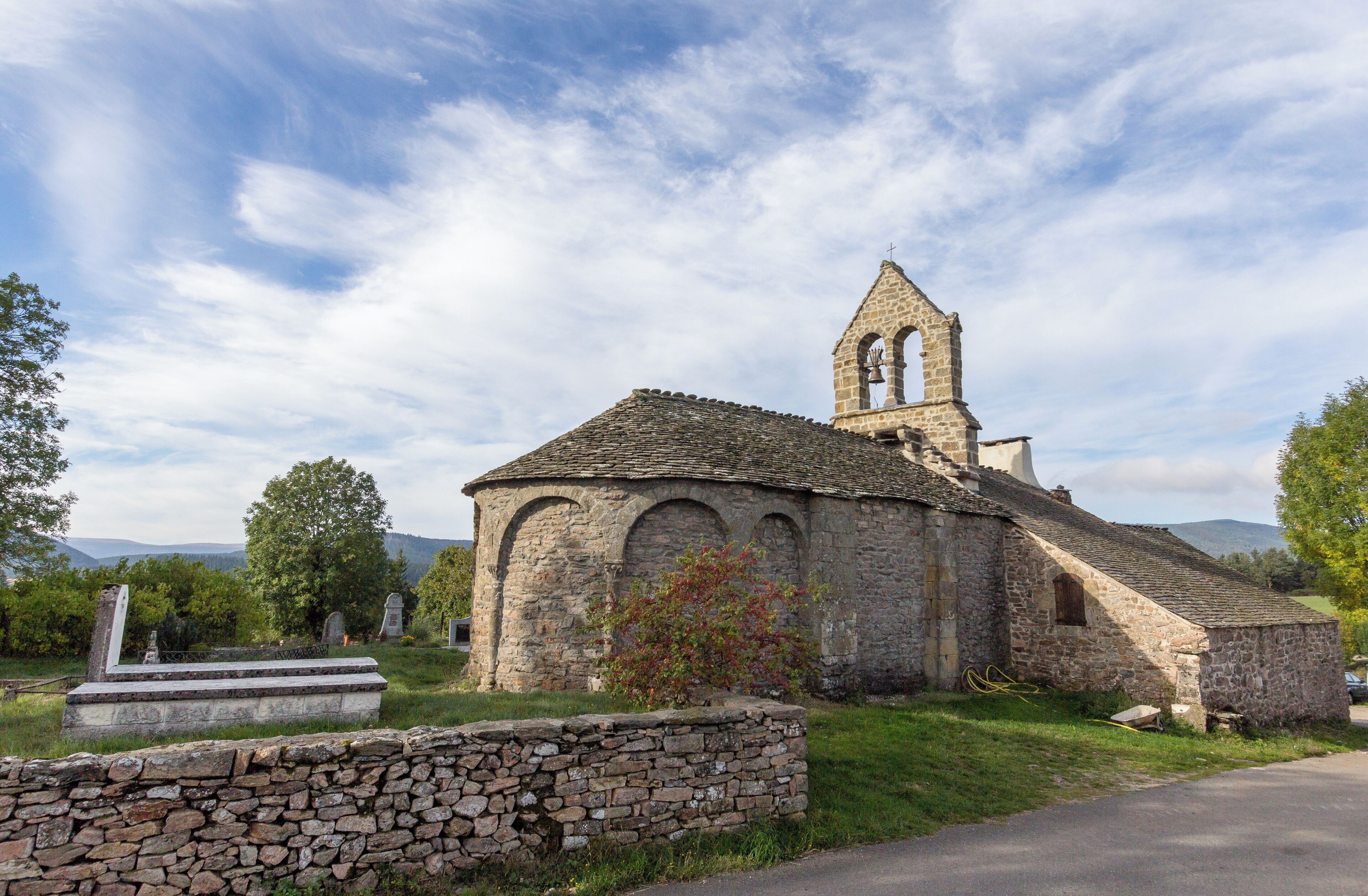A stone church with a bell tower and arched windows, surrounded by greenery and a stone wall.