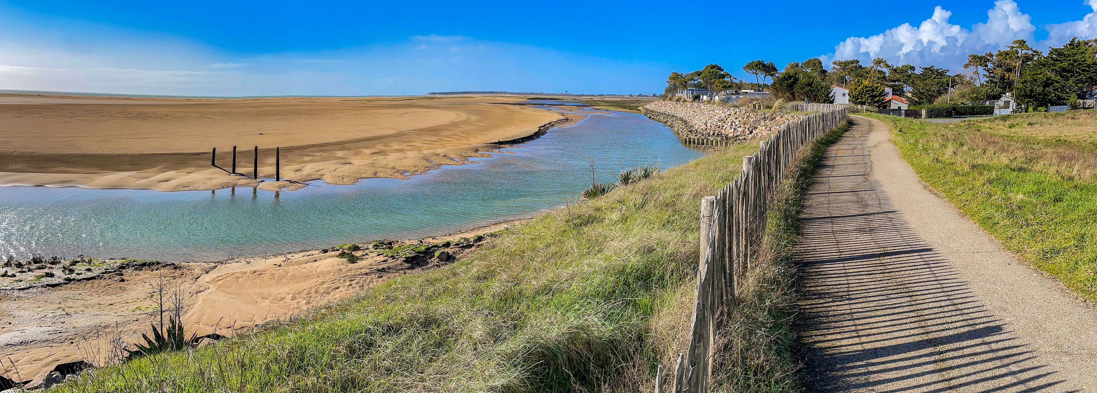 A coastal landscape with a sandy beach, a narrow water channel, and a dirt path alongside a wooden fence.
