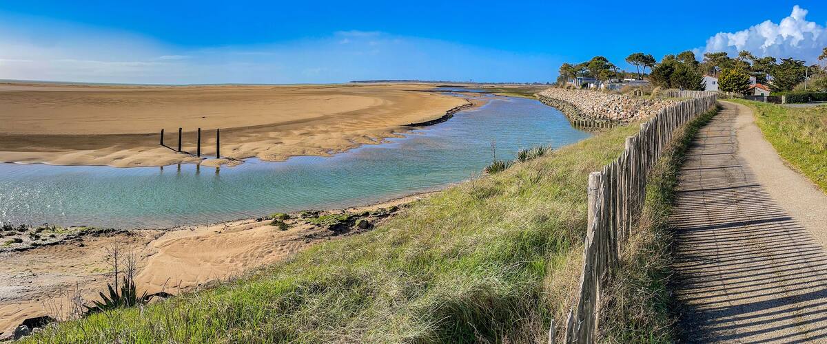 A coastal landscape with a sandy beach, a narrow water channel, and a dirt path alongside a wooden fence.