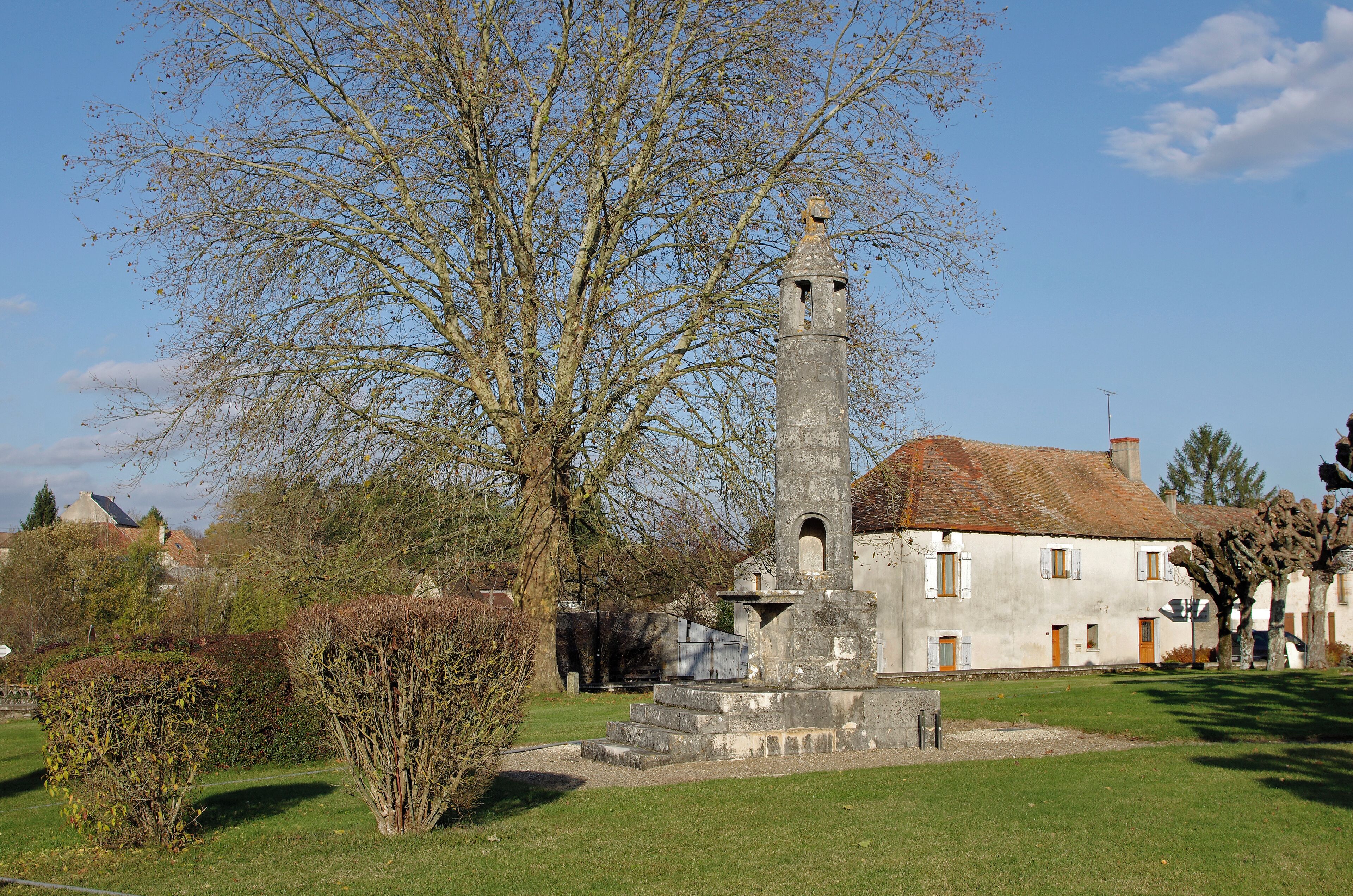 A historic tower in a village setting with a large tree in the foreground and a white building with a red roof in the background.