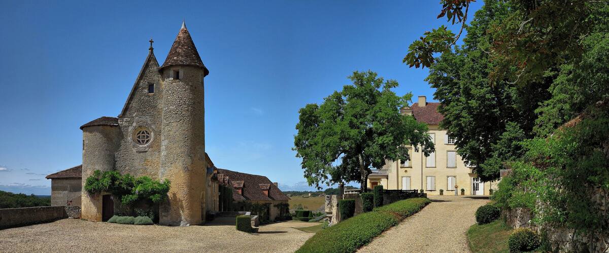 A historic stone tower and a large building with a tiled roof, surrounded by a gravel courtyard and lush greenery.