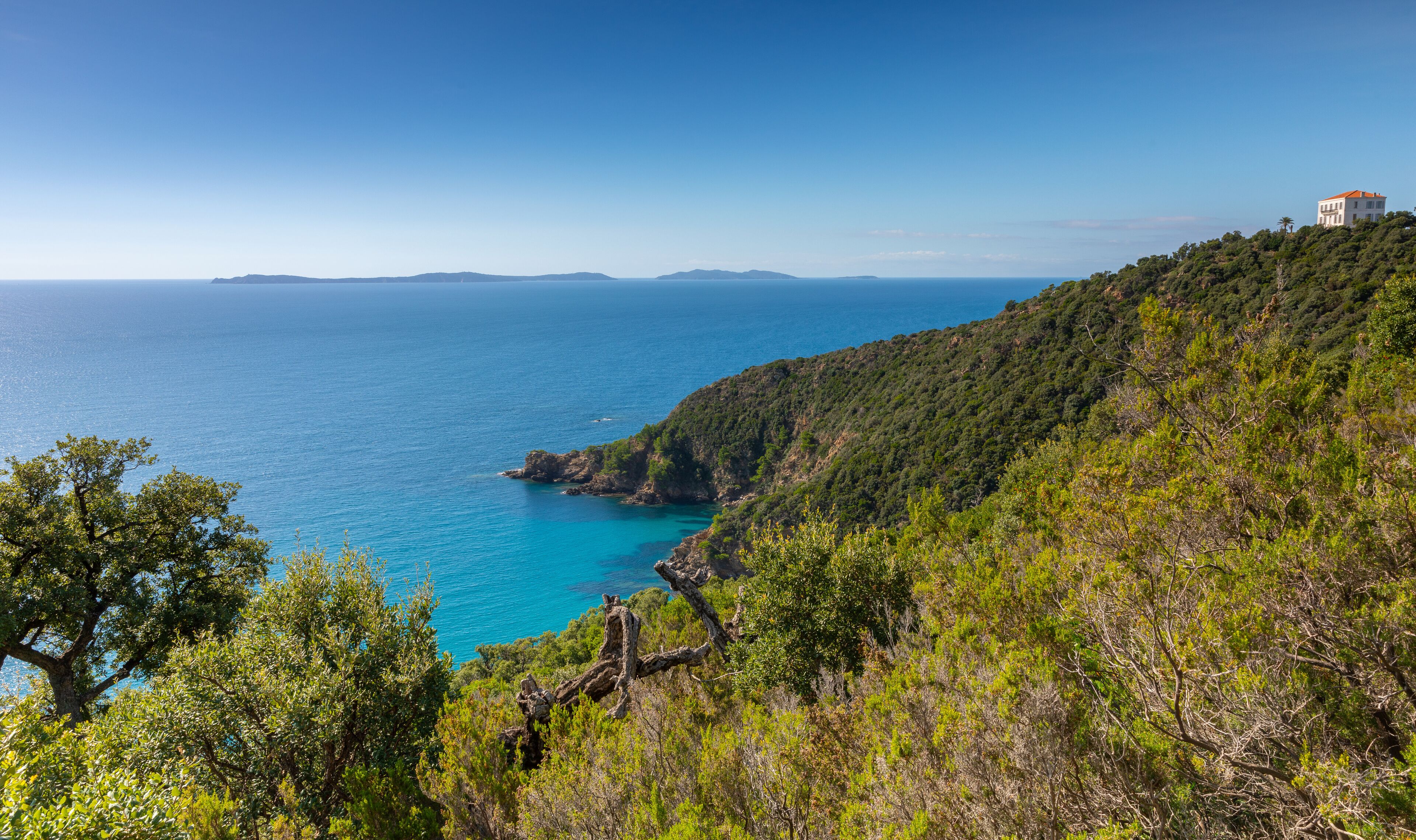 A coastal landscape with a clear blue sea, green hills, and a white building on a hill.