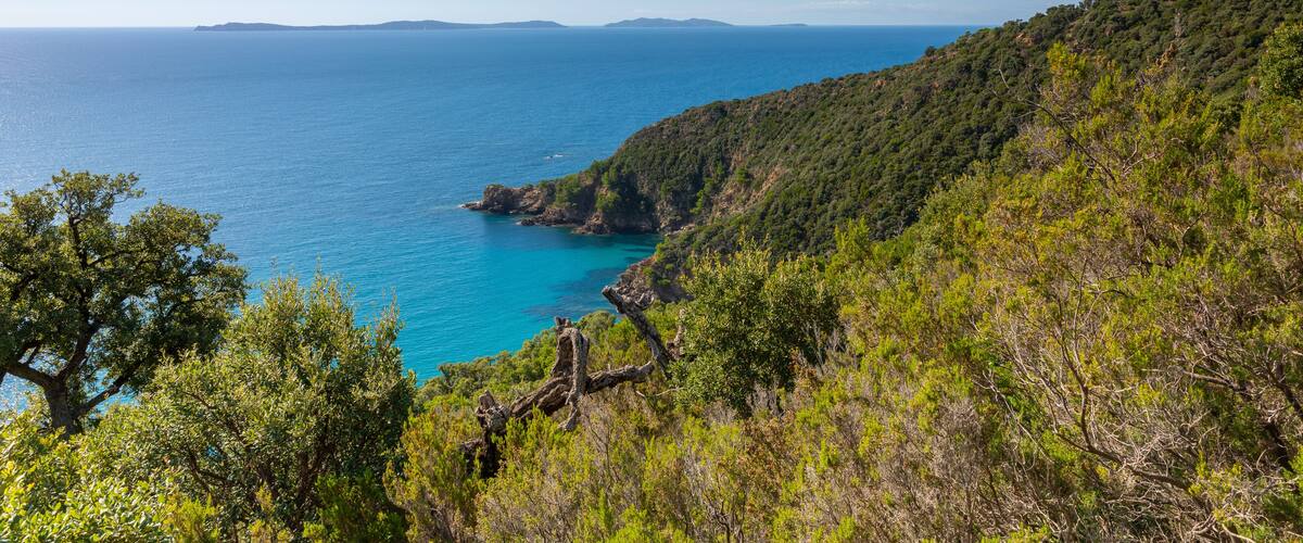 A coastal landscape with a clear blue sea, green hills, and a white building on a hill.