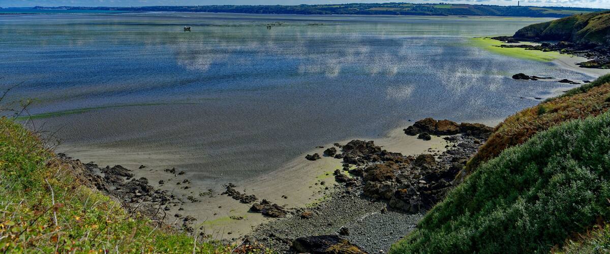 A coastal landscape with a sandy beach, rocky outcrops, and a distant shoreline under a partly cloudy sky.
