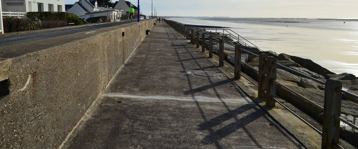 A coastal promenade with a concrete walkway, wooden railings, and houses along the shore.