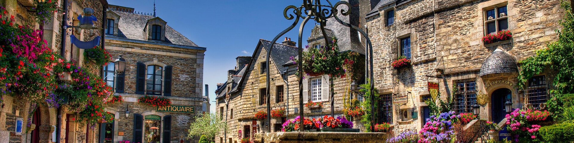 Una calle adoquinada con edificios históricos de piedra, macetas con flores y una fuente en el centro.