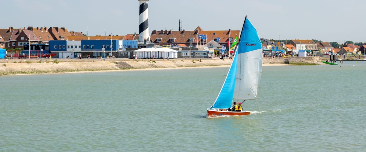 A sailboat with a blue sail on a calm body of water, a lighthouse, and buildings along the shore.