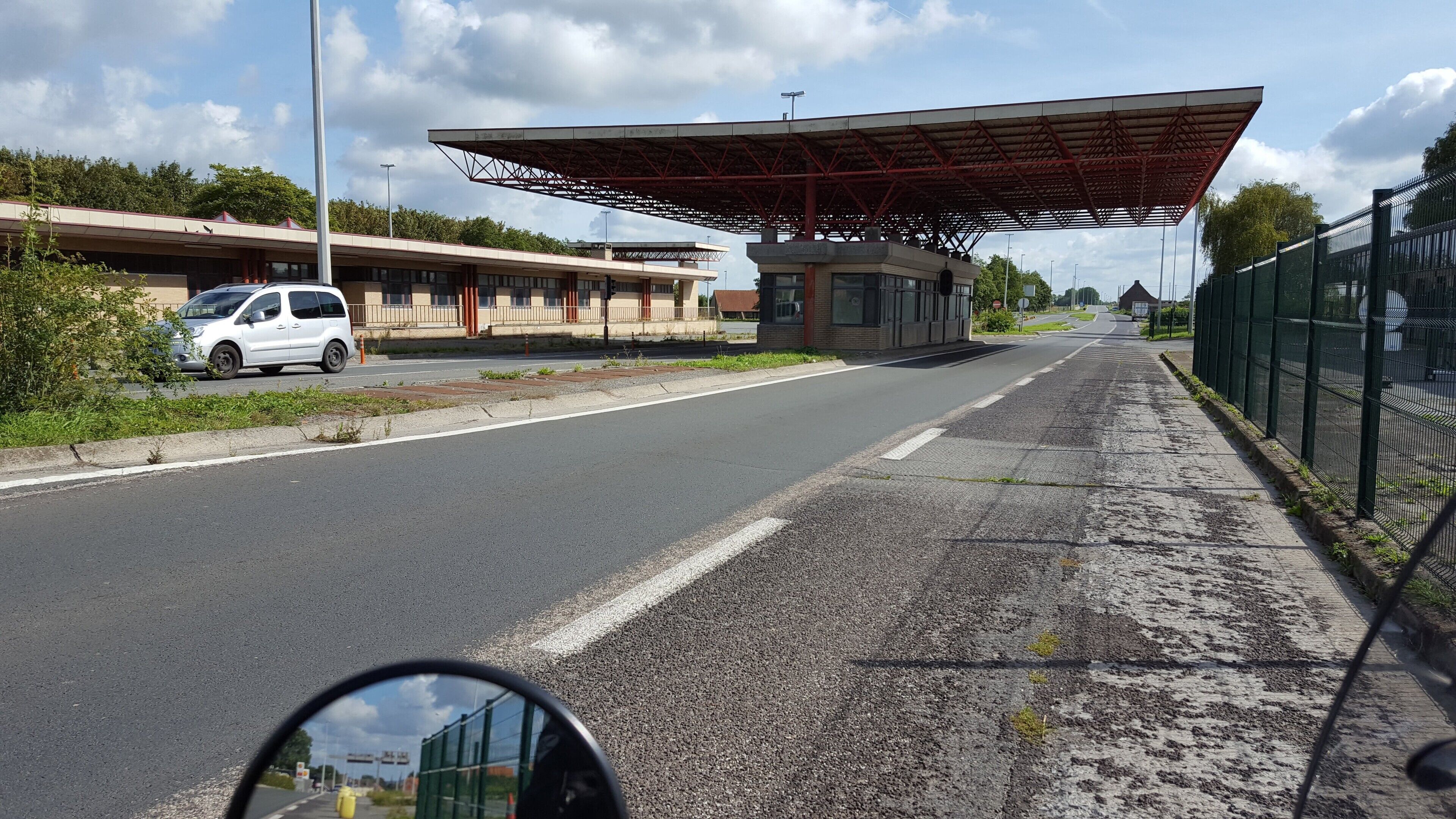 A road with a white car parked on the side, a covered structure, and a green fence.