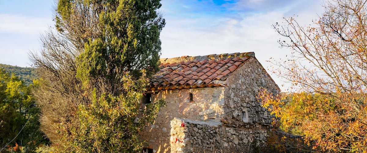 A stone building with a red-tiled roof surrounded by tall grass and trees.