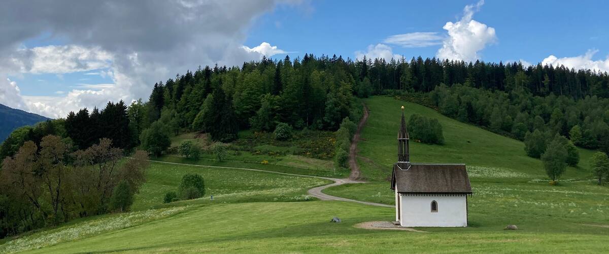 A white church on a grassy hill with a forested background and a partly cloudy sky.