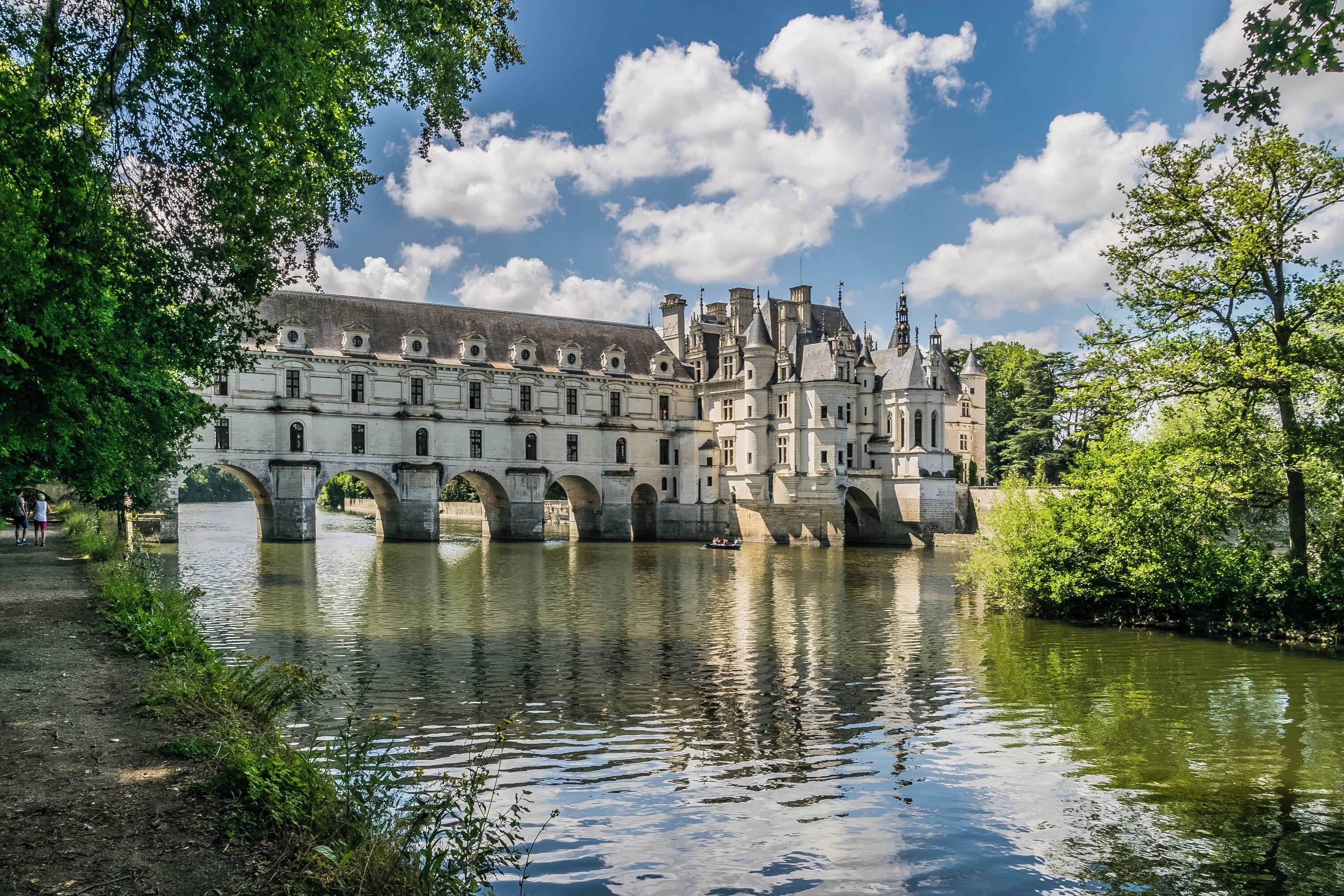 A historic building with a river in front, featuring a bridge and lush greenery.