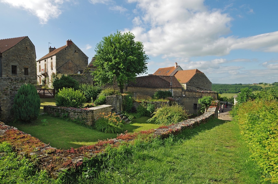 Un village historique aux bâtiments en pierre, bordé d’une allée d’arbres et offrant un paysage verdoyant luxuriant.