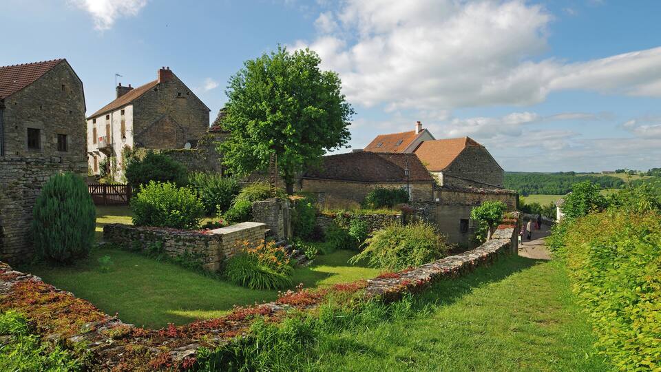 A historic village with stone buildings, a tree-lined pathway, and a lush green landscape.