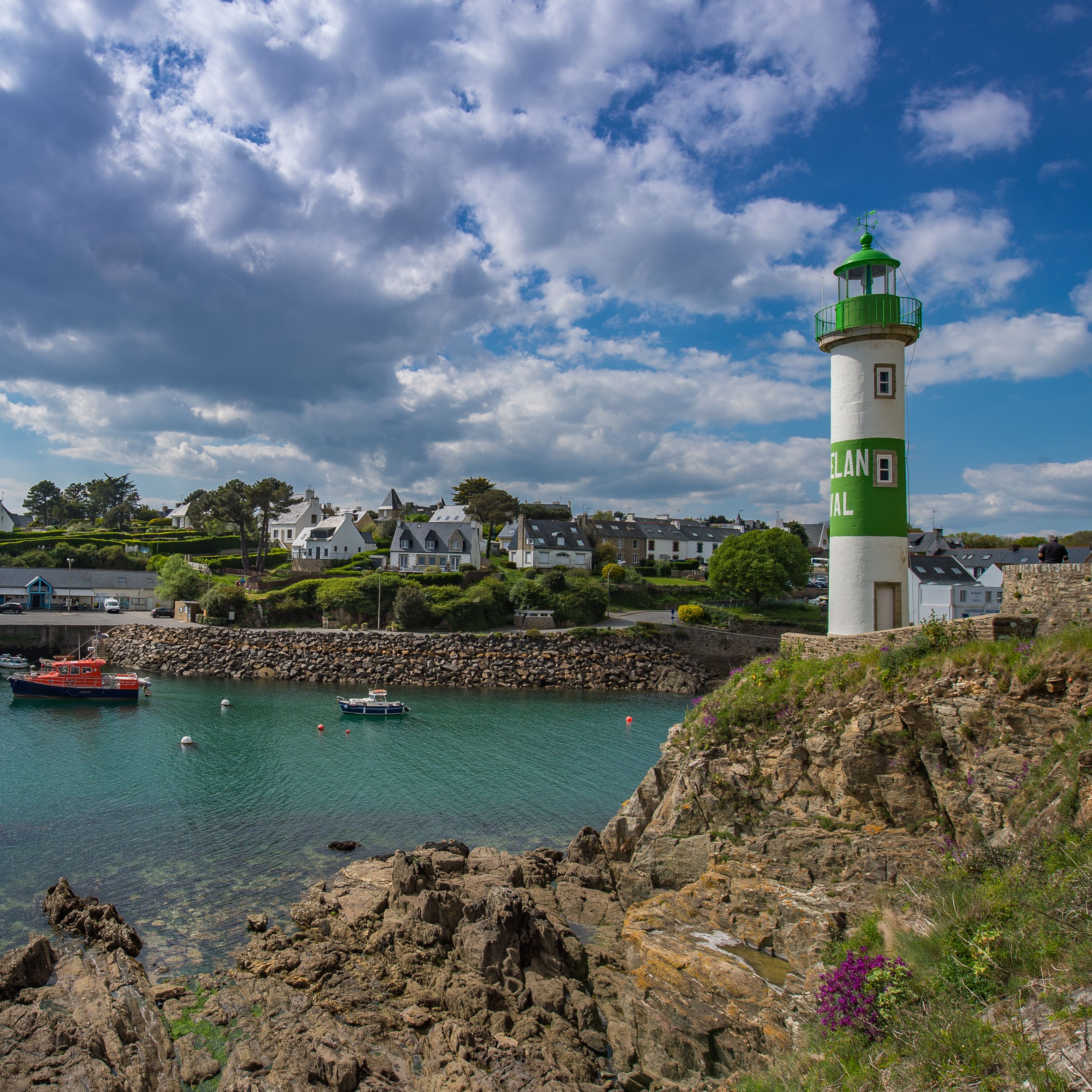A coastal town with a prominent green and white lighthouse, calm waters, and rocky cliffs.