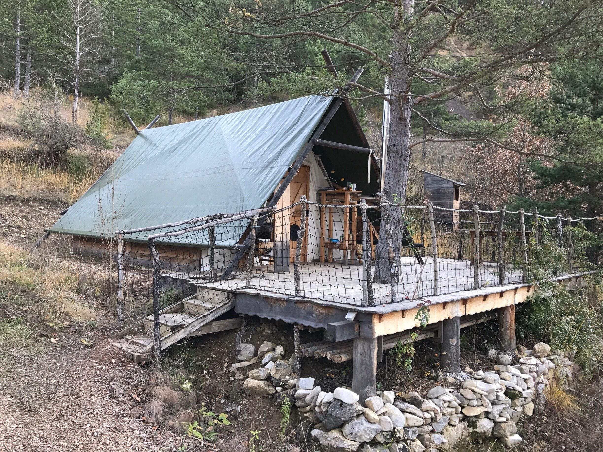 A rustic cabin with a green roof, wooden deck, and stone walls, surrounded by trees and a stone foundation.