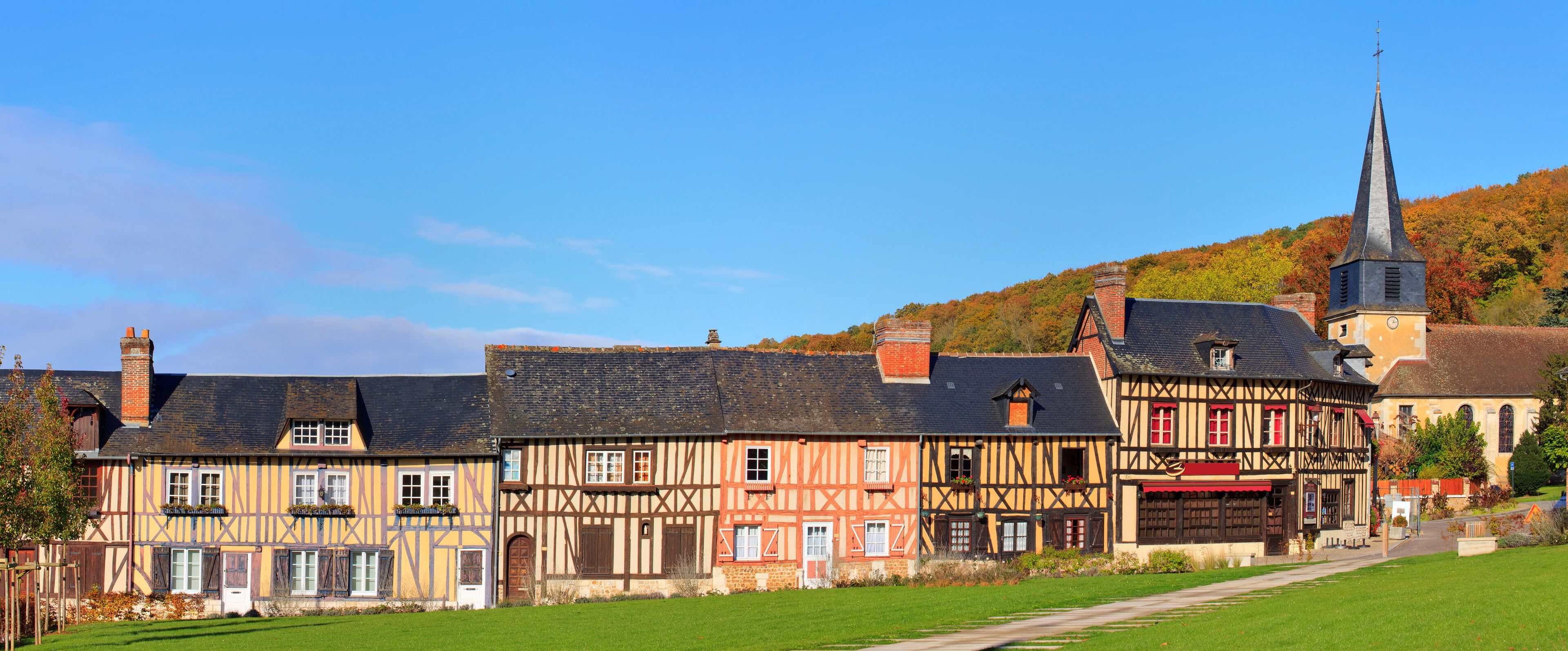 A picturesque village with traditional half-timbered houses, a church with a tall steeple, and a lush green hillside in the background.