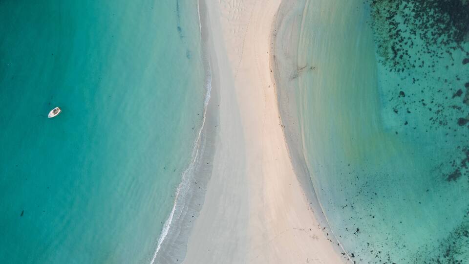 An aerial view of a beach with clear turquoise water, a sandy shoreline, and a small boat.