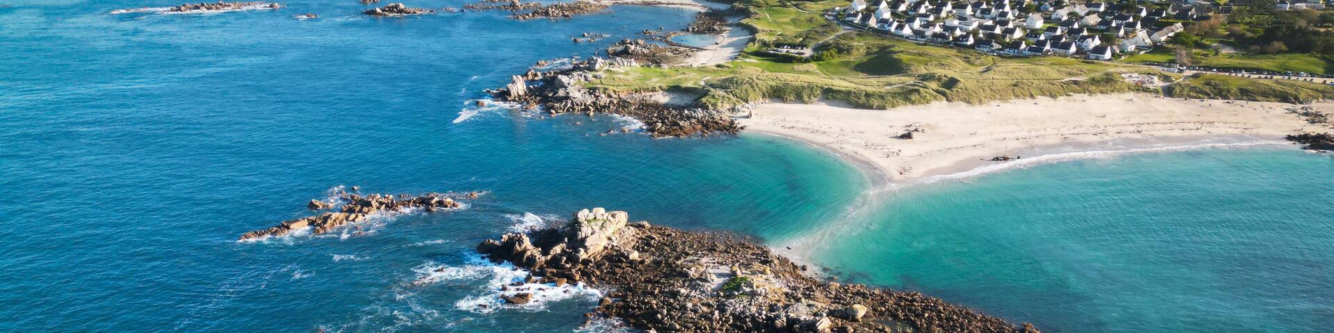 Aerial view of the rugged coastline where dark rocks meet the turquoise sea, contrasting with the green fields, Cleder, France.