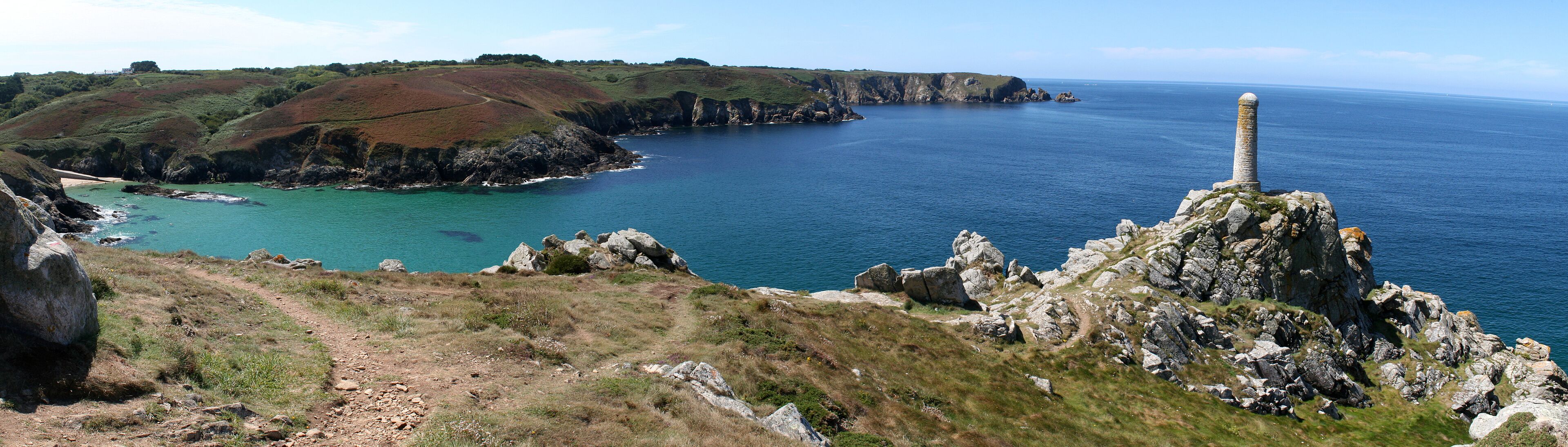 A coastal landscape with rocky cliffs, a clear blue sea, and a prominent stone monument.