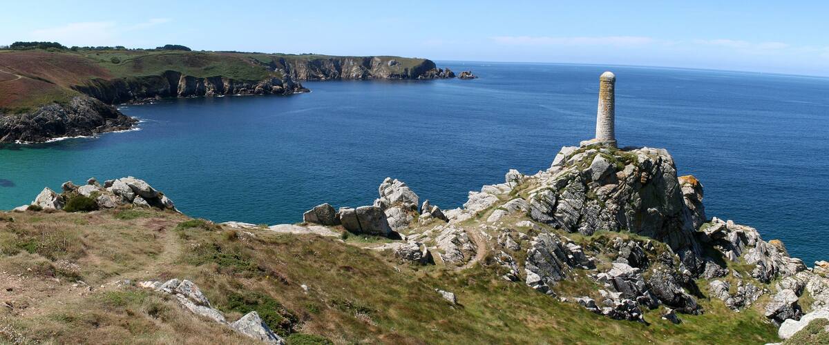 A coastal landscape with rocky cliffs, a clear blue sea, and a prominent stone monument.