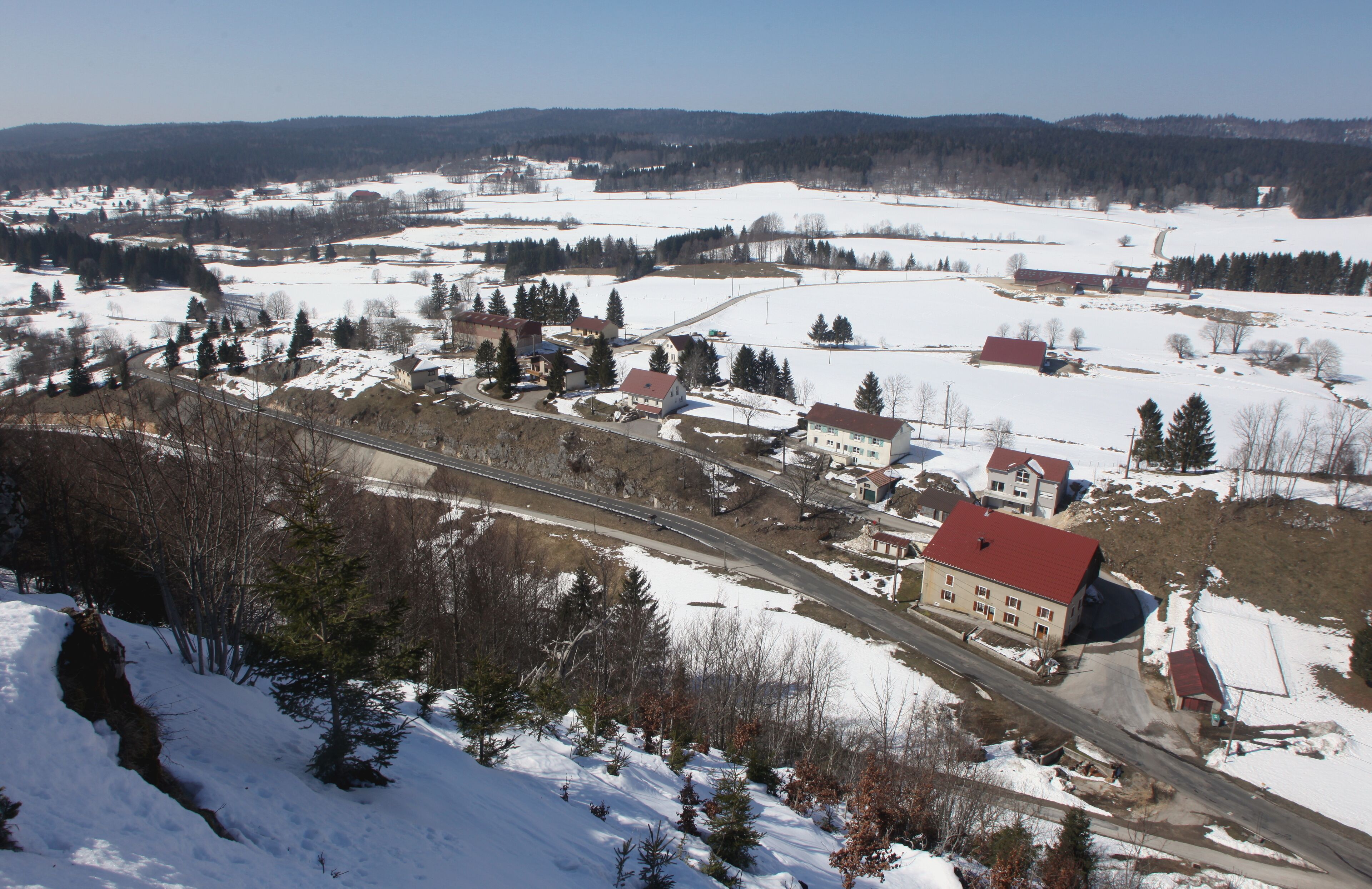 A snow-covered landscape with a railway track, buildings, and a forested area.