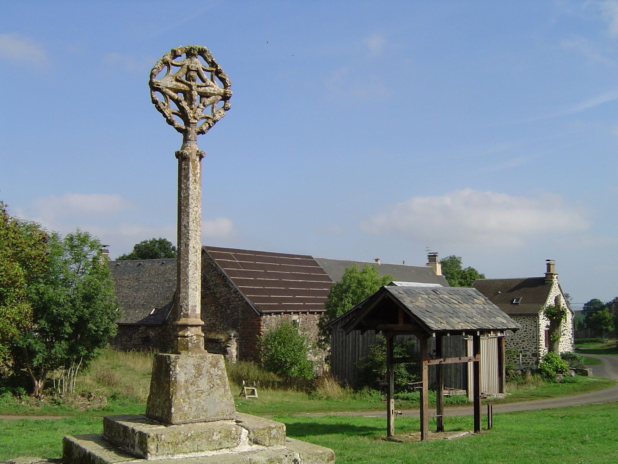 A stone monument with a circular design, a stone building with a sloped roof, and a small wooden structure in a rural setting.