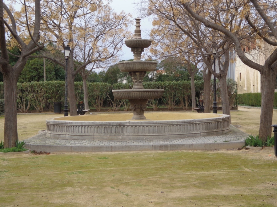 Una fuente de piedra de varios niveles en un parque rodeado de árboles y un edificio al fondo.