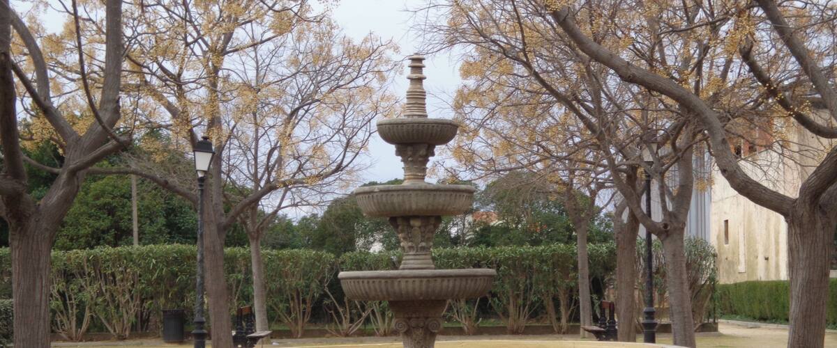 A stone fountain with multiple tiers in a park surrounded by trees and a building in the background.