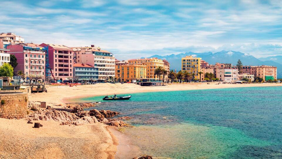 A coastal cityscape with a sandy beach, colorful buildings, and mountains in the background.