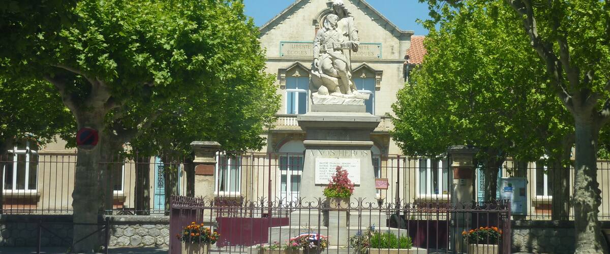A building with a statue of a soldier on a pedestal, surrounded by trees and a fence with flower boxes.